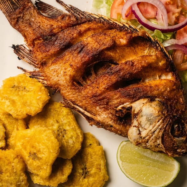 Whole fried fish served with golden plantain slices, lime wedge, and lettuce, tomato, and red onion salad.