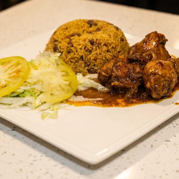 Plate of saucy braised chicken, seasoned rice, shredded lettuce, and sliced green tomatoes on a countertop.
