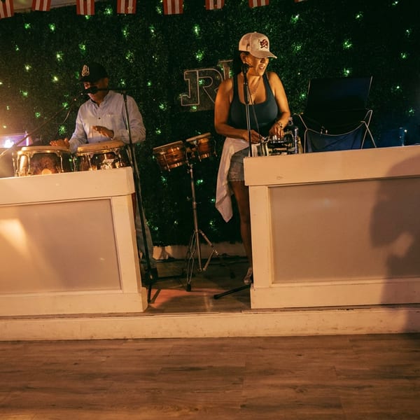 Two musicians perform percussion and DJ music on a warmly lit restaurant stage with a sparkling green backdrop.