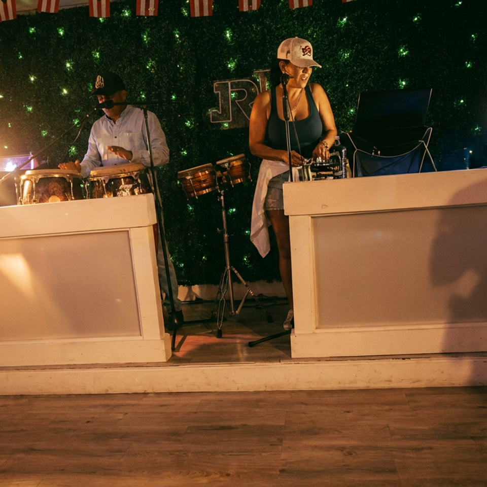 Two musicians perform percussion and DJ music on a warmly lit restaurant stage with a sparkling green backdrop.