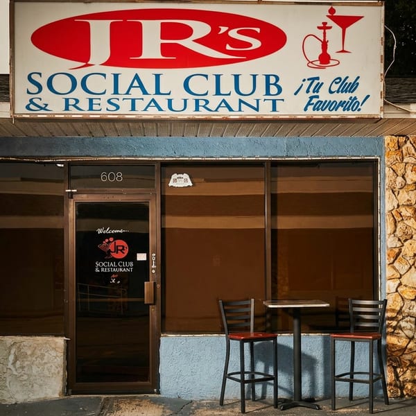 Exterior of JR’s Social Club & Restaurant with stone facade, entrance, outdoor high-top table, and sign advertising lunch specials.