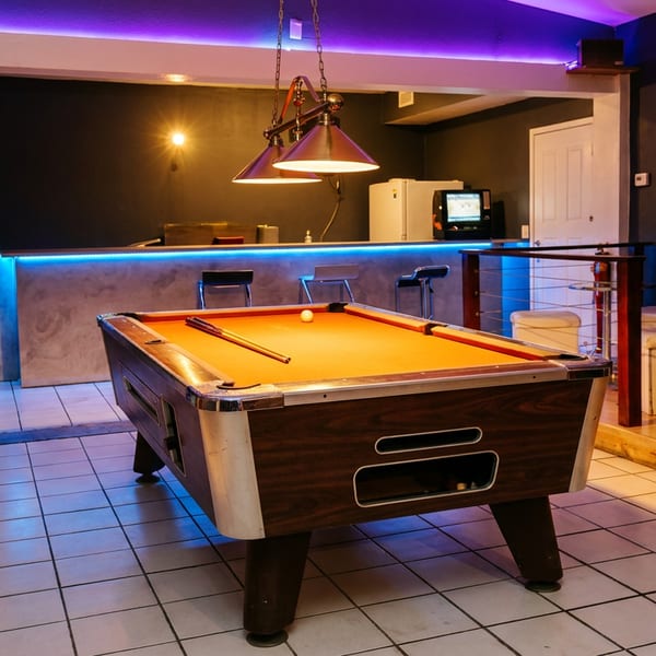 Pool table with cue sticks in a neon-lit bar lounge featuring a blue-lit counter, stools, and seating area.
