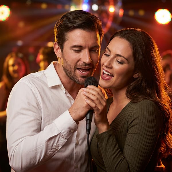 Couple singing together into a microphone at a lively, warmly lit restaurant karaoke event with dancing guests.