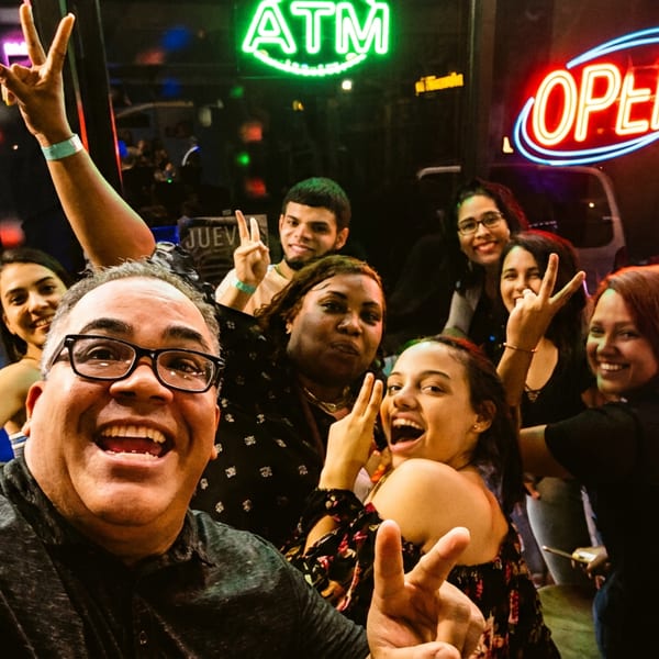 Group of smiling friends posing with peace signs outside a lively restaurant beneath illuminated ATM and OPEN signs.
