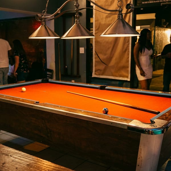 Pool table with orange felt and cue beneath pendant lights in a lively restaurant or bar.