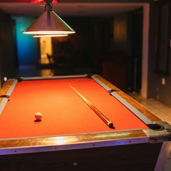 Red-felt pool table with cue and billiard ball beneath a hanging lamp in a dimly lit restaurant game room.