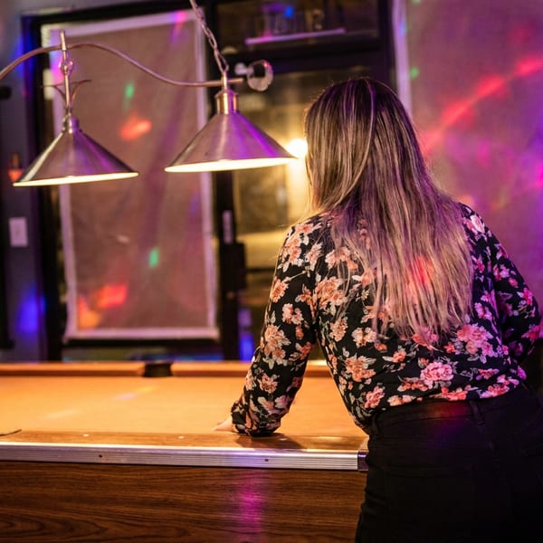 Woman in a floral blouse leans over a pool table beneath colorful nightclub lights.