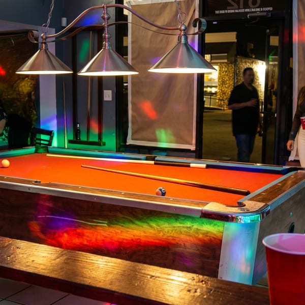 Pool table with orange felt, billiard balls, and cue under pendant lights in a colorful restaurant entertainment area.