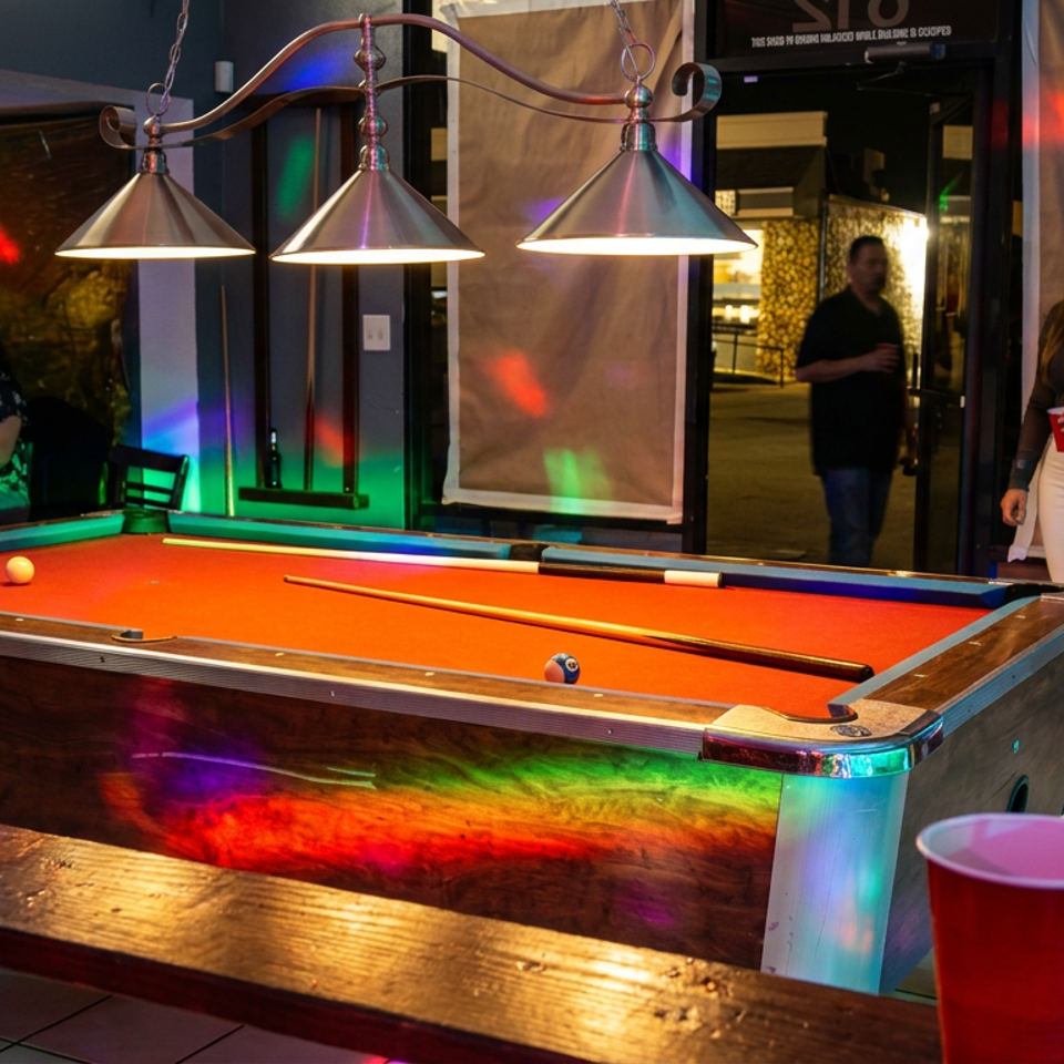 Pool table with orange felt, billiard balls, and cue under pendant lights in a colorful restaurant entertainment area.